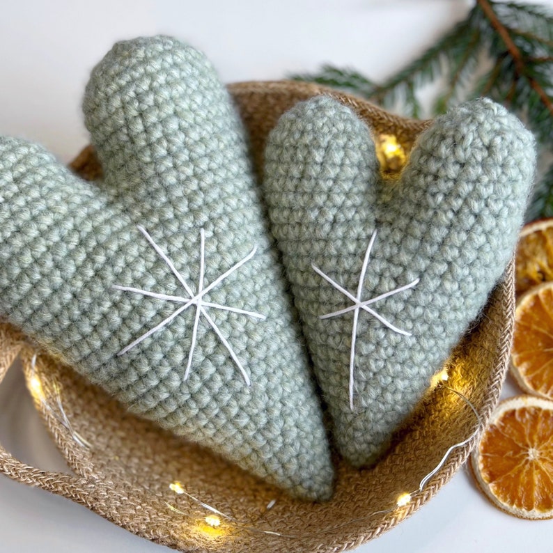 May include: Two crocheted green heart-shaped ornaments with white snowflake designs. The hearts are sitting in a woven basket with string lights.