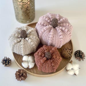 Op de afbeelding: Drie handgemaakte, gebreide pompoenen in beige, poederroze en roestkleuren, elk met een glinsterende bruine steel. De pompoenen zijn gerangschikt op een houten dienblad met katoenbollen en dennenappels, wat een herfstthema decoratie creëert.