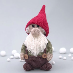 May include: A crocheted gnome with a red hat, white beard, green and brown clothing, and brown button accents.