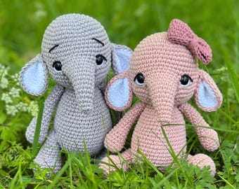 Puede incluir: Dos peluches de elefante de crochet, uno gris y otro rosa, sentados en la hierba verde. El elefante rosa tiene un pequeño lazo rosa en la cabeza.