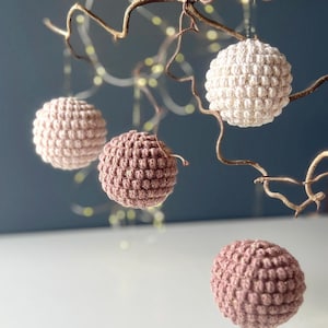 May include: Four crocheted spheres in varying shades of pink and cream hang from slender, brown branches. The spheres have a textured, bobble-like surface. The background is a muted teal, with soft, out-of-focus lights adding a touch of sparkle. These are likely decorative ornaments.