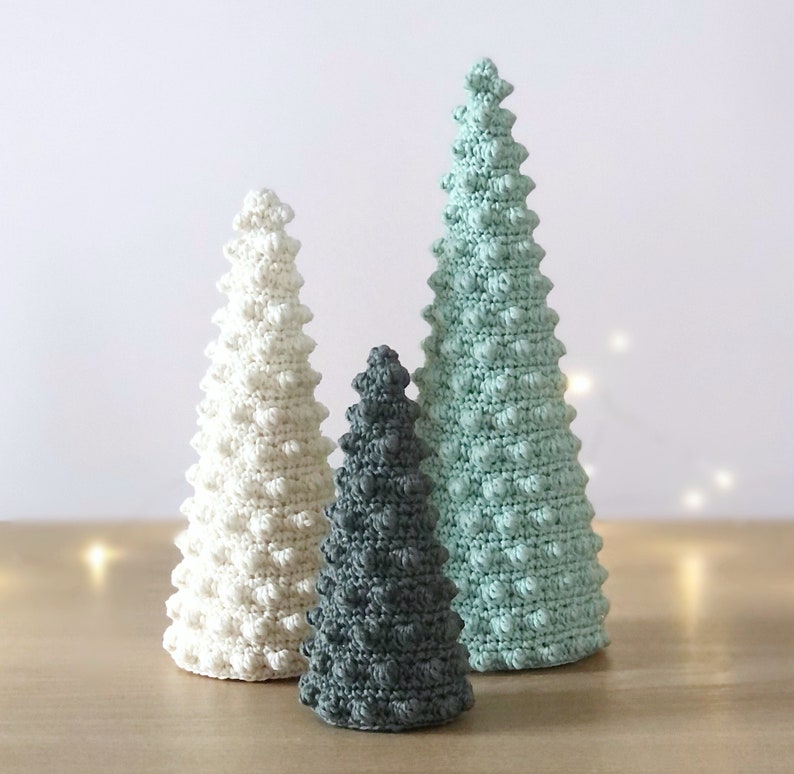 May include: Three crocheted Christmas trees in different sizes. The largest tree is light green, the middle tree is white, and the smallest tree is gray.