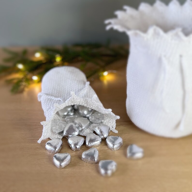 May include: A white crocheted drawstring bag filled with silver foil-wrapped heart-shaped chocolates. The bag is spilling out the chocolates onto a wooden surface.