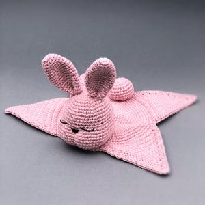 以下が含まれることがあります： 星型のデザインのピンクの編みぐるみうさぎのセキュリティブランケット。うさぎは小さな丸い尻尾と目を閉じている。