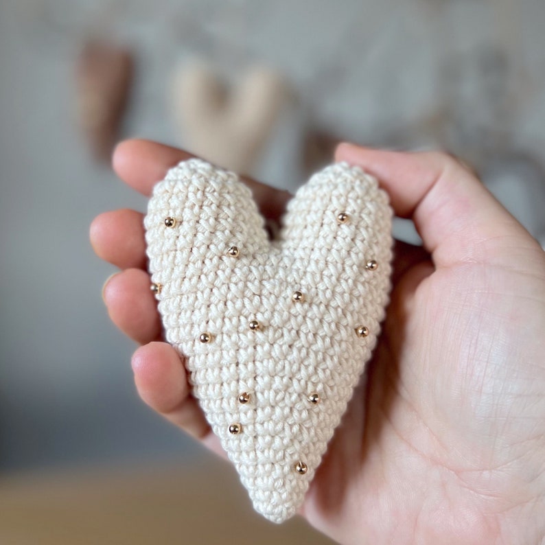 May include: A crocheted heart-shaped ornament with a white background and gold beads scattered across the surface.