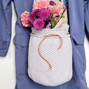 以下が含まれることがあります： 薄いグレーのクロシェのトートバッグに、茶色の革のストラップとピンクと紫の花束が飾られています。