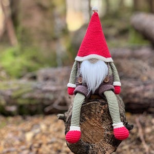 Op de afbeelding: Een gehaakte gnome met een rode muts, witte baard en bruine overall zit op een boomstronk in een bos.