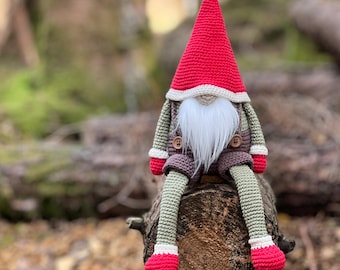 May include: A crocheted gnome with a red hat, white beard, and brown overalls sits on a tree stump in a forest setting.
