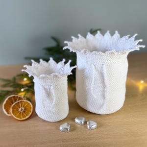 May include: Two white crocheted baskets with scalloped edges, the larger one is taller than the smaller one. Three silver foil-wrapped heart-shaped chocolates are in front of the baskets.