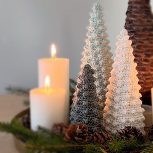 K&ouml;nnte beinhalten: Drei geh&auml;kelte Weihnachtsb&auml;ume in Wei&szlig;, Grau und Blau, mit einer wei&szlig;en Kerze im Vordergrund. Die B&auml;ume sind mit kleinen wei&szlig;en und grauen Kugeln verziert.