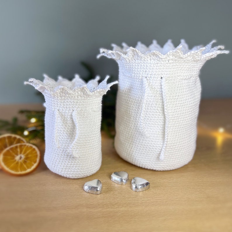 May include: Two white crocheted drawstring bags with scalloped edges. The larger bag is in the background and the smaller bag is in the foreground. There are three silver foil-wrapped heart-shaped chocolates in the foreground.