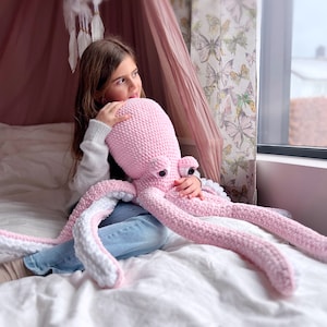 Puede incluir: Una niña pequeña abraza un gran peluche de pulpo de crochet rosa. El pulpo tiene detalles blancos en sus tentáculos y grandes ojos amigables.