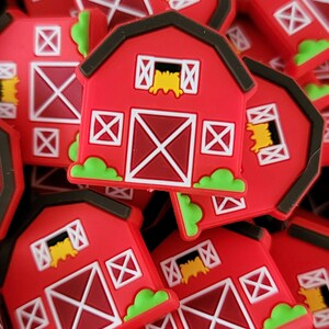 May include: A pile of red barn-shaped charms with white accents and green grass. The charms have a hay bale in the window and a white X pattern on the door.