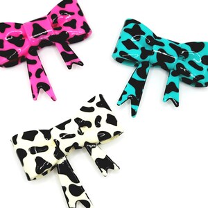 May include: Three decorative bow hair clips in vibrant colors. One is hot pink with black spots, another is turquoise with black spots, and the third is white with black spots. The bows have a glossy finish and are arranged on a white background.