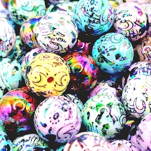 May include: A close-up shot of a pile of colorful, round beads. The beads are various colors, including blue, yellow, pink, and iridescent. Each bead has a unique design in black, with swirls and other patterns. The beads are likely for jewelry making.