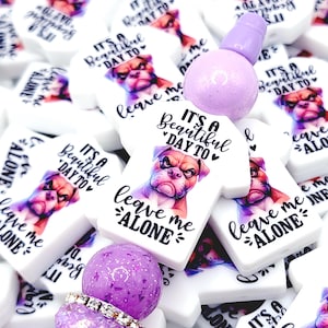 May include: A collection of white rectangular beads with a bulldog graphic and the text "IT'S A Beautiful DAY TO leave me ALONE". A purple bead and a purple bottle stopper are also visible.