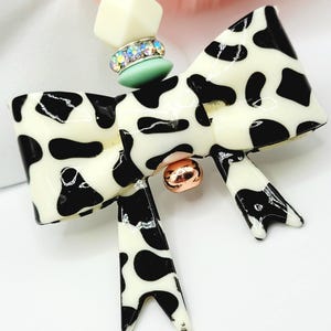May include: A cow-print bow pendant necklace with a cream-colored base and black spots. The bow has a gold-colored center and is connected to a beaded necklace with pink, cream, and green beads. The necklace is a fashion accessory.