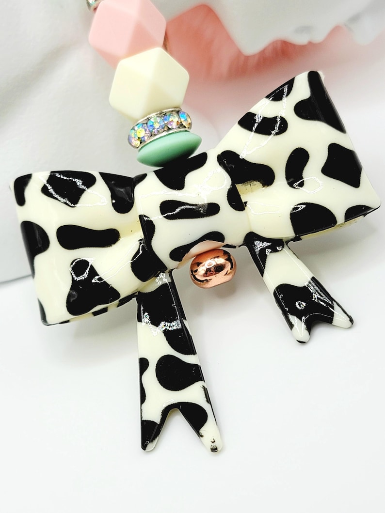 May include: A cow-print bow pendant necklace with a cream-colored base and black spots. The bow has a gold-colored center and is connected to a beaded necklace with pink, cream, and green beads. The necklace is a fashion accessory.