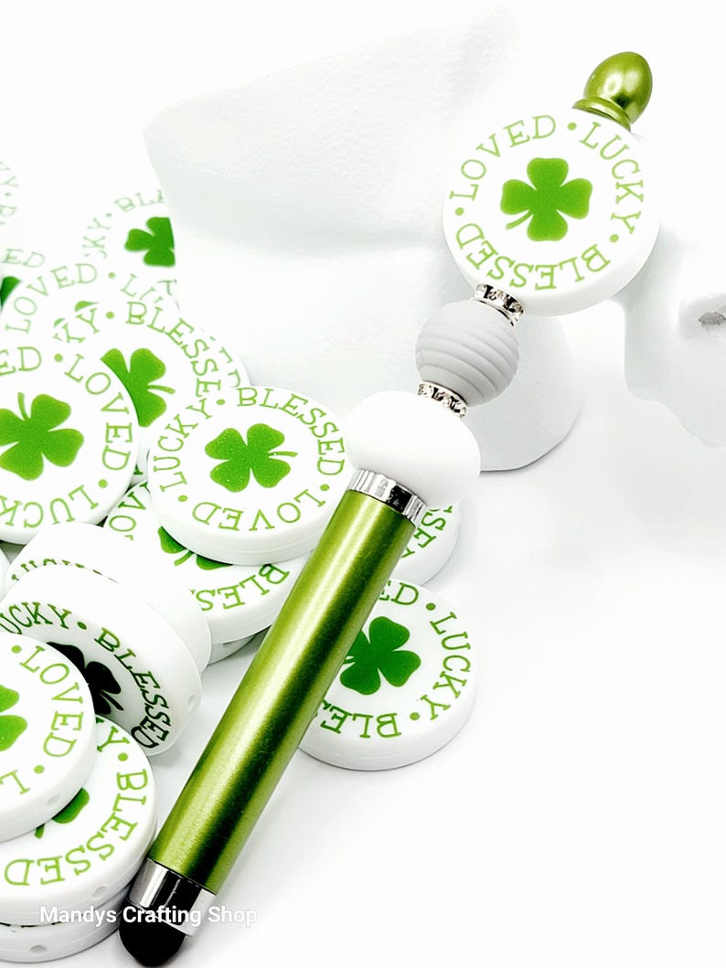 Peut inclure: Stylet vert et blanc avec un charme rond blanc qui dit "Blessed. Loved. Lucky." avec un tr&egrave;fle &agrave; quatre feuilles vert au centre. Le charme est attach&eacute; &agrave; une perle grise et une perle blanche.