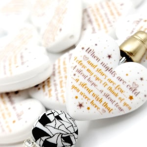 May include: White heart-shaped beads with gold text and star accents. The text reads "When nights are cold, I close my eyes & think of you...a silent wish that you were here." A black and white patterned bead is also visible.