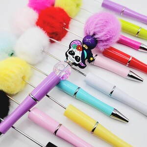 May include: A collection of colourful pens with fluffy pom-poms on the top. The pens are in various colours, including pink, yellow, red, white, blue, green, and black. One pen has a skull charm with a witch's hat and flowers. The pens are arranged in a row on a white surface.