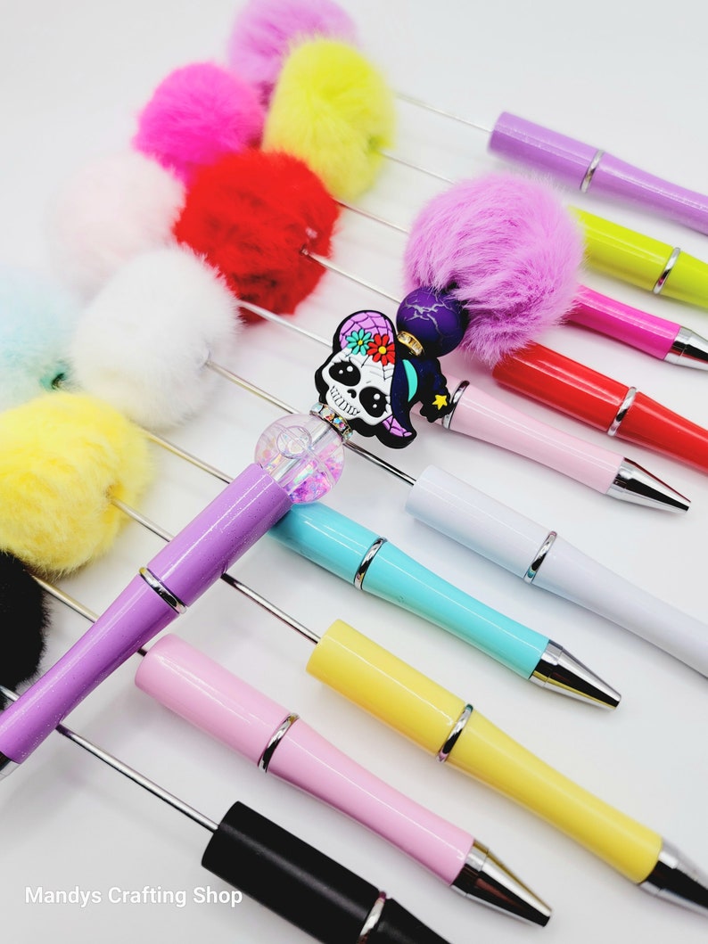 May include: A collection of colourful pens with fluffy pom-poms on the top. The pens are in various colours, including pink, yellow, red, white, blue, green, and black. One pen has a skull charm with a witch's hat and flowers. The pens are arranged in a row on a white surface.