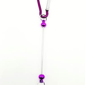 Op de afbeelding: Paarse metalen sleutelhanger met een karabijnhaak, een metalen draad en twee paarse metalen ballen. De draad is bevestigd aan de karabijnhaak en de ballen.
