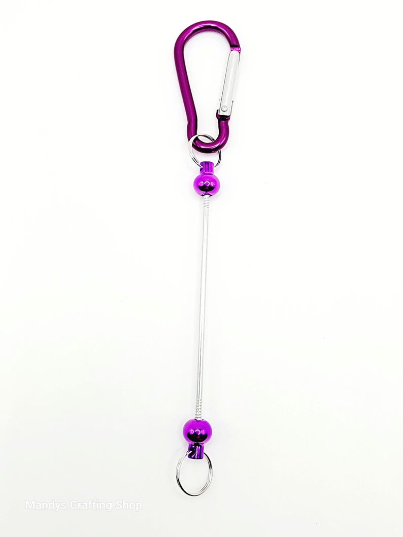 Op de afbeelding: Paarse metalen sleutelhanger met een karabijnhaak, een metalen draad en twee paarse metalen ballen. De draad is bevestigd aan de karabijnhaak en de ballen.