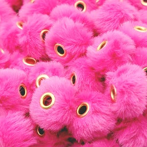 May include: Close-up of a pile of bright pink, fluffy pom-poms with gold eyelets. The pom-poms are densely packed together, creating a textured surface. The eyelets are evenly spaced and add a touch of metallic contrast to the soft, plush material.
