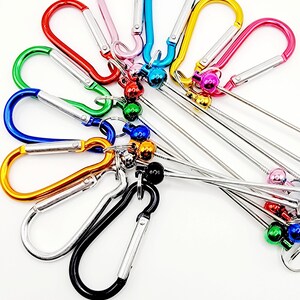 Op de afbeelding: Assortiment van kleurrijke metalen karabijnhaken met bevestigde metalen ringen en belletjes. De karabijnhaken zijn in verschillende kleuren, waaronder roze, blauw, groen, rood, oranje, zilver en zwart.