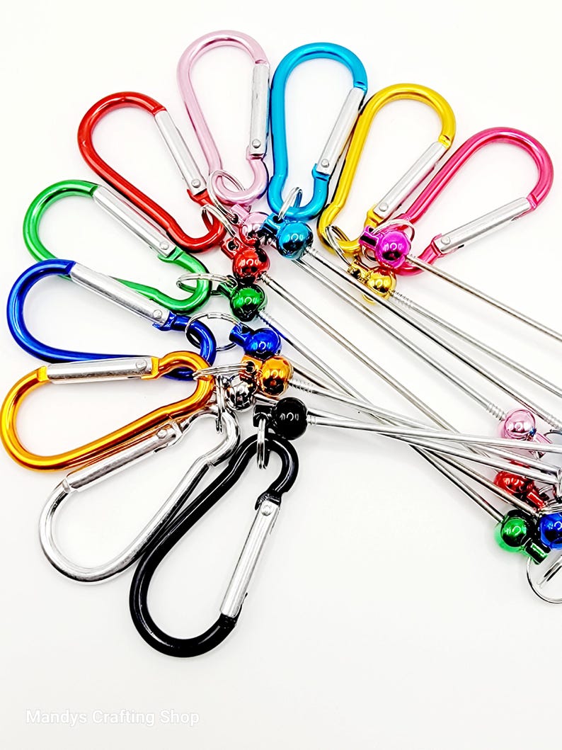 Op de afbeelding: Assortiment van kleurrijke metalen karabijnhaken met bevestigde metalen ringen en belletjes. De karabijnhaken zijn in verschillende kleuren, waaronder roze, blauw, groen, rood, oranje, zilver en zwart.