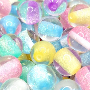May include: A collection of colorful, transparent glass marbles. The marbles feature a variety of pastel colors, including pink, blue, yellow, and purple, with glitter accents. Each marble has a unique design, with some featuring stripes and others a swirl pattern.