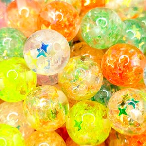 May include: A close-up shot of a collection of colorful, transparent glass beads. The beads are in shades of orange, green, yellow, and clear, with star-shaped and crescent moon-shaped designs inside. The beads are scattered, creating a vibrant and playful display.