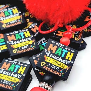 May include: Black rectangular keychain with the text "I'M A MATH Teacher OF COURSE I HAVE PROBLEMS" in white, yellow, and blue. The keychain has a red fluffy pom-pom and a red keychain ring. The theme is math teacher gifts.