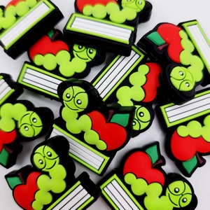 May include: A group of green and red book charms with a worm and apple design. The charms are made of rubber and have a black outline.