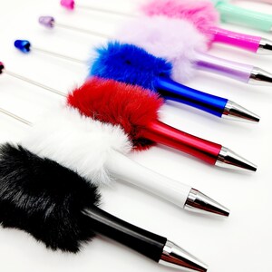 May include: A set of ten colourful pens with fluffy pom-poms on the top. The pens are arranged in a row, with the pom-poms facing the viewer. The colours of the pom-poms are black, white, red, blue, purple, pink, light blue, green, and pink.