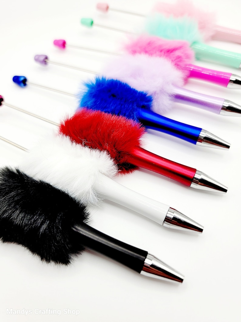 May include: A set of ten colourful pens with fluffy pom-poms on the top. The pens are arranged in a row, with the pom-poms facing the viewer. The colours of the pom-poms are black, white, red, blue, purple, pink, light blue, green, and pink.
