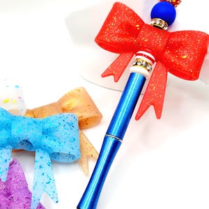 May include: A blue pen with a red glitter bow and a decorative top. The pen has a silver tip and is surrounded by other colorful bow-shaped accessories. The pen's design includes a blue bead and a gold and red beaded accent.