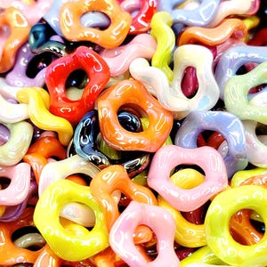 May include: A close-up shot of a pile of colorful, glossy beads in various shapes. The beads are in shades of red, orange, yellow, pink, green, blue, and purple. The shapes include rings and curved, organic forms. The beads are likely for jewelry making or crafts.