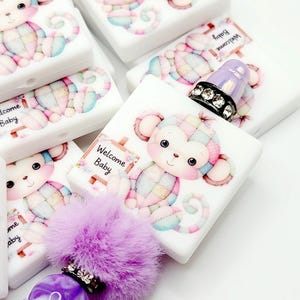 May include: A purple and white monkey charm with a patchwork design and a sign that says "Welcome Baby". The charm is attached to a purple pom-pom and a purple and white bead.