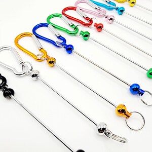 Op de afbeelding: Assortiment van kleurrijke metalen sleutelhangers met een karabijnhaak en een bal aan het einde van een metalen ketting. De sleutelhangers zijn verkrijgbaar in zwart, oranje, geel, groen, blauw, rood, roze, lichtblauw en paars.