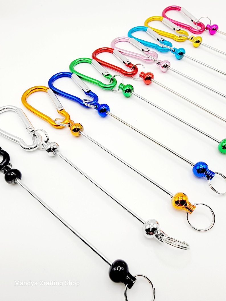 Op de afbeelding: Assortiment van kleurrijke metalen sleutelhangers met een karabijnhaak en een bal aan het einde van een metalen ketting. De sleutelhangers zijn verkrijgbaar in zwart, oranje, geel, groen, blauw, rood, roze, lichtblauw en paars.