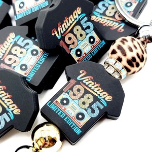 May include: A black and gold keychain with a vintage 1985 t-shirt design. The keychain features a cassette tape and the text "Vintage 1985 Limited Edition".