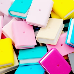 May include: A pile of colorful miniature book charms in various colors, including pink, blue, yellow, and white. The charms have a small hole in the center for attaching to a keychain or other items.