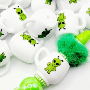 May include: A collection of white ceramic mugs with green alien illustrations. The mugs are arranged in a pile, with a green fuzzy pom-pom on top. The mugs are perfect for adding a fun and quirky touch to any kitchen or office.