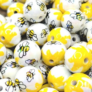 May include: A collection of round beads with a bee theme. The beads are white with yellow accents and feature a black and yellow bee design. Some beads are solid yellow. These beads are suitable for jewelry making and crafts.