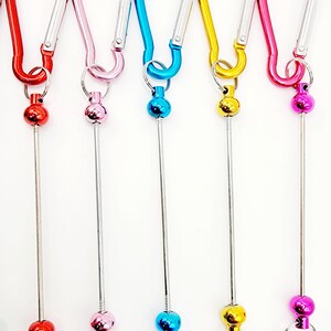 Op de afbeelding: Zes kleurrijke metalen sleutelhangers met een draaibare bal en een karabijnhaak. De sleutelhangers zijn rood, roze, blauw, geel en twee tinten roze.