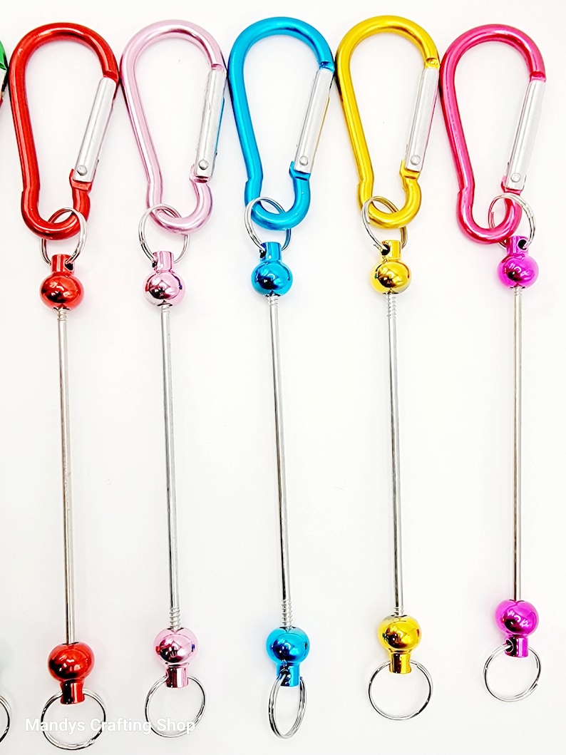 Op de afbeelding: Zes kleurrijke metalen sleutelhangers met een draaibare bal en een karabijnhaak. De sleutelhangers zijn rood, roze, blauw, geel en twee tinten roze.