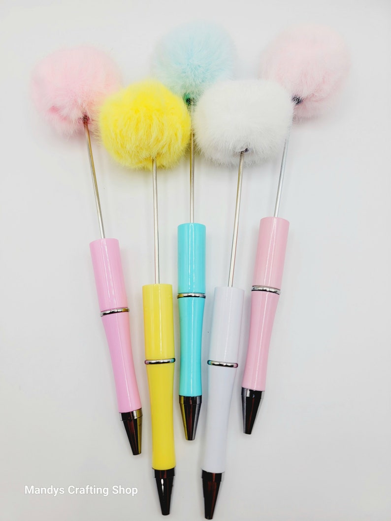 May include: Five colourful ballpoint pens with fluffy pom-poms on top. The pens are pink, yellow, blue, white, and light pink.