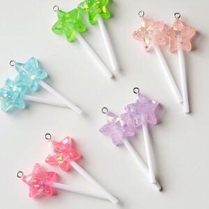 May include: A collection of colorful star-shaped charms with white wands. The charms are in shades of green, blue, pink, and purple, with iridescent glitter accents. Each charm has a silver loop at the top for attaching to jewelry or other crafts.